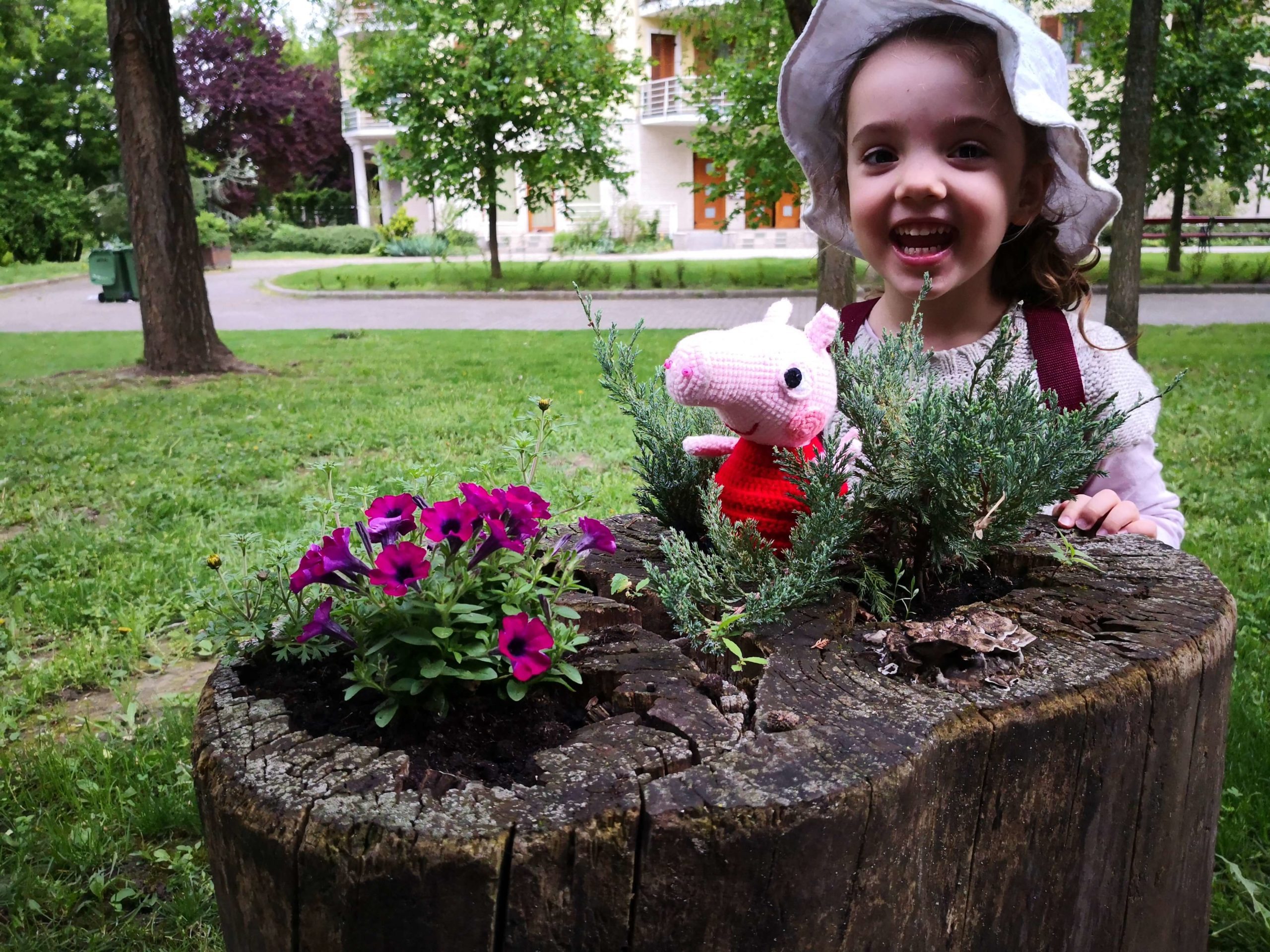 Kind hält selbstgehäkelten Peppa-Wutz-Kopf in der Hand, im Garten bei Blumen – fröhlicher Moment beim Häkeln