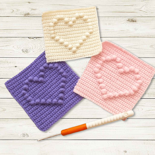 Gehäkelte Granny Squares mit Herzmotiv in Weiß, Rosa und Violett auf Holzuntergrund