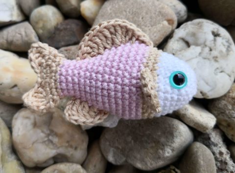 Gehäkelter kleiner Fisch in Rosa und Beige mit Sicherheitsauge, dekorativ auf Steinen fotografiert