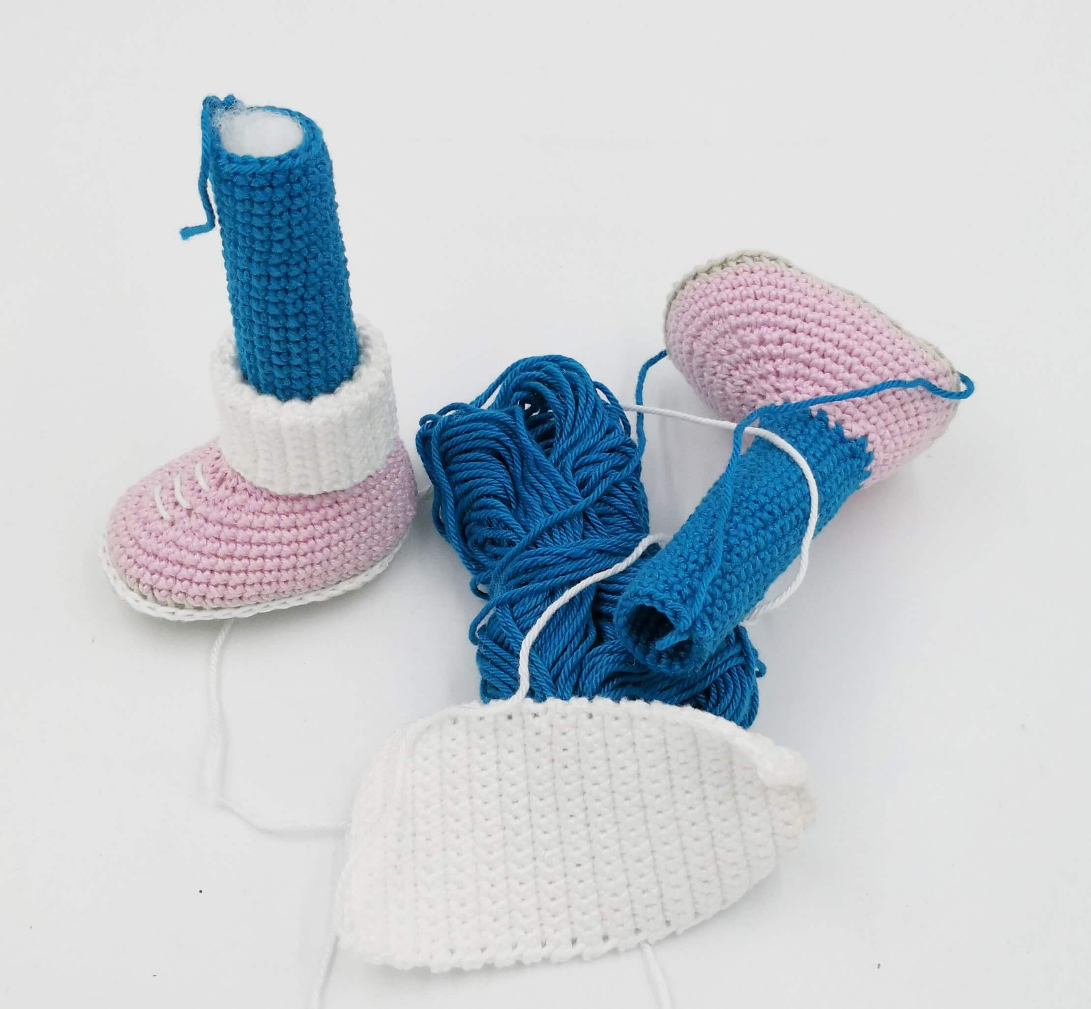 Gehäkelte Beine einer Amigurumi-Puppe mit weißen Stiefeln, blauen Hosen und rosa Schuhsohlen – noch nicht zusammengenäht