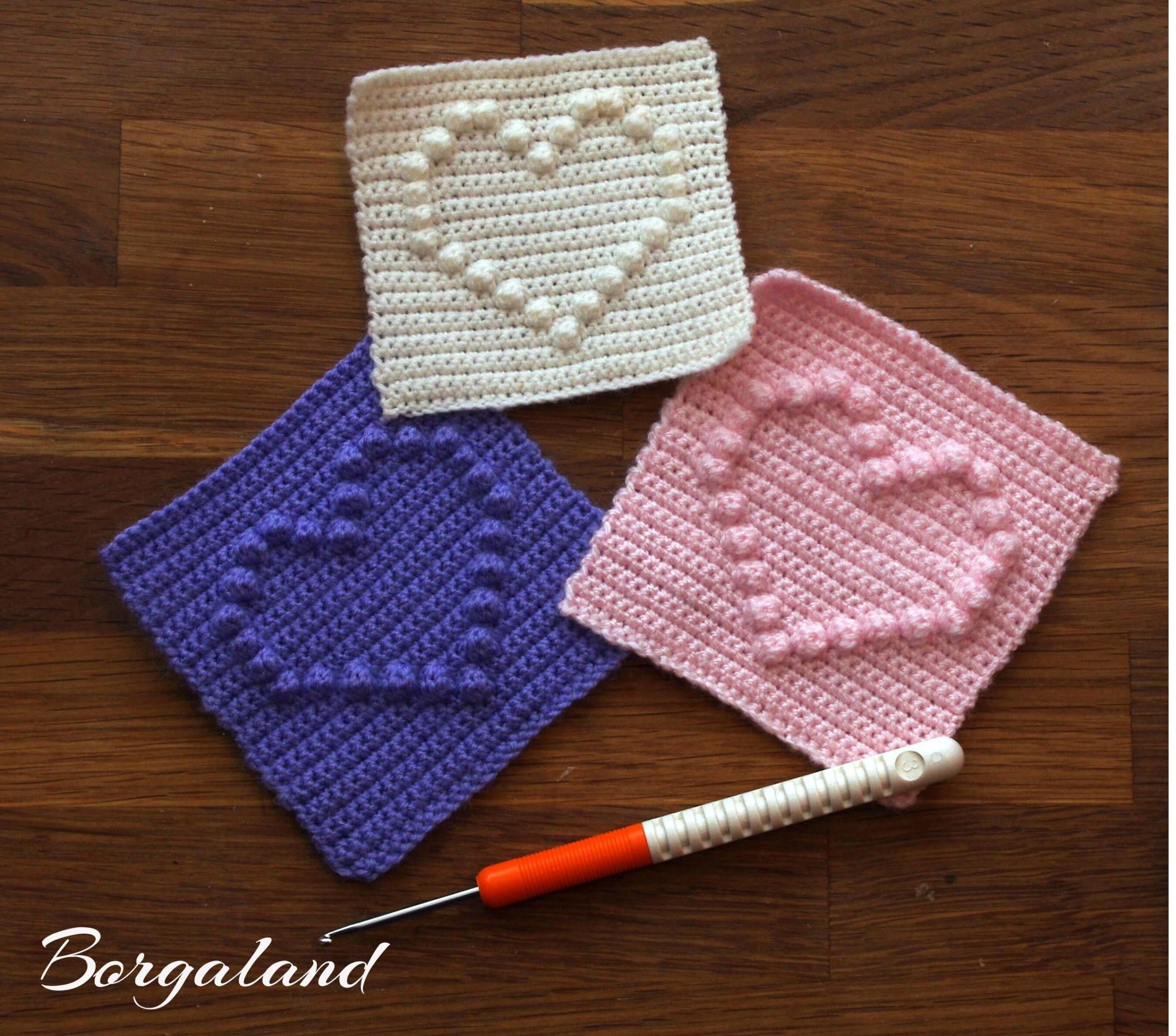 Gehäkelte Granny Squares mit Herzmotiv in Weiß, Rosa und Violett auf Holzuntergrund