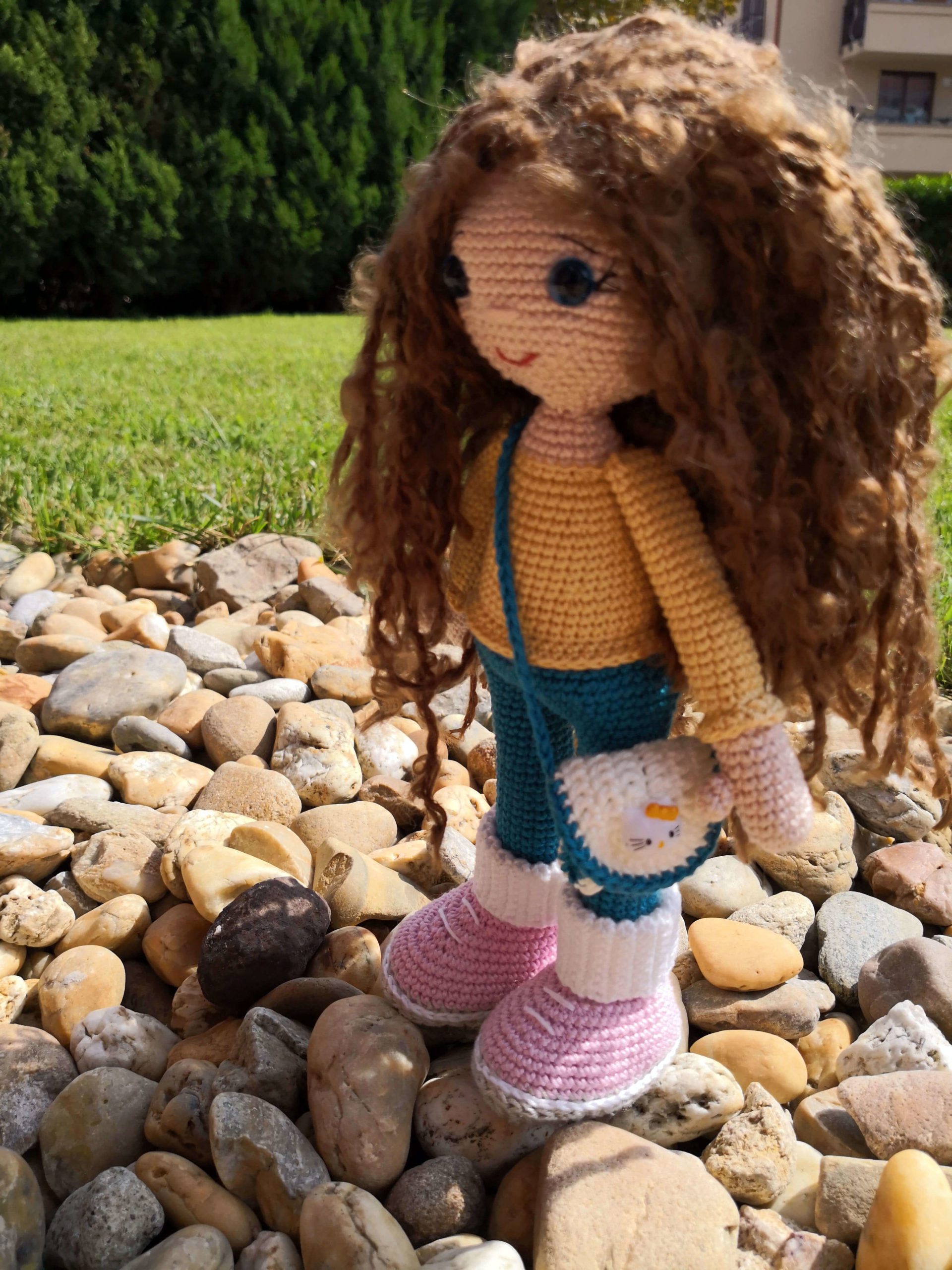 Gehäkelte Amigurumi-Puppe mit lockigem Haar, Jeans, gelbem Pullover und Umhängetasche – draußen auf Steinweg fotografiert