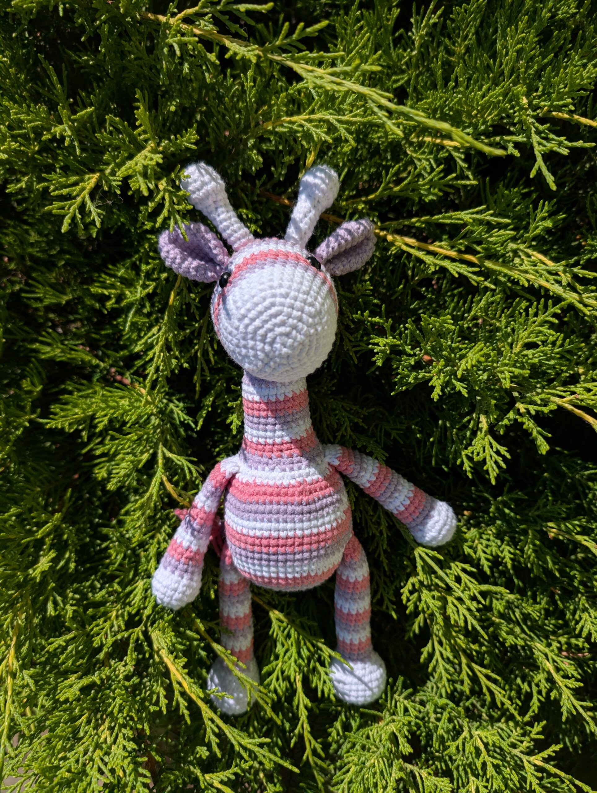 Frontalansicht einer gehäkelten Amigurumi-Giraffe mit rosa-lila Streifen, fotografiert im Grünen.